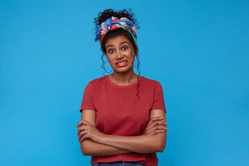 Bewildered young pretty brunette curly woman with gathered hair showing her teeth while looking confusedlly at camera, isolated against blue background