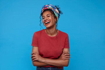 Happy young lovely dark haired woman with gathered hair throwing back her head while laughing happily, standing over blue background with folded hands