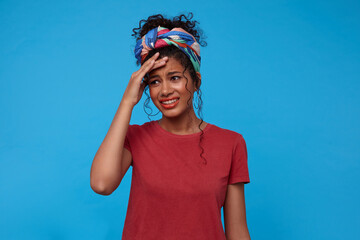 Indoor photo of young pretty dark haired curly lady with festive makeup frowning her face and keeping raised hand on forehead while posing over blue background