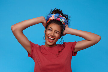 Joyful young lovely dark haired woman with casual hairstyle winking happily with broad smile and holding raised hands on her head, isolated over blue background