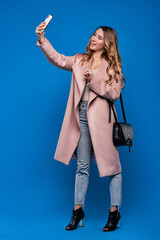 .Young beautiful blonde girl in a raincoat on a blue background with mobile phone doing selfie