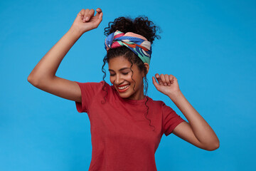 Good looking young pretty brunette curly woman with gathered hair smiling cheerfully with closed eyes while dancing with raised hands, posing over blue background