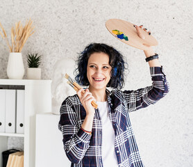 Smiling woman artist in her studio holding art palette and paintbrushes