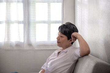 Happy Middle-aged gray-haired Asian woman wearing a white shirt poses at home on a sunny morning, a single retired woman sitting on the sofa in the living room. Positive single senior retired Concept