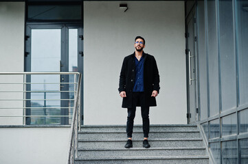 Middle eastern entrepreneur wear black coat and blue shirt, eyeglasses against office building, stand on stairs.