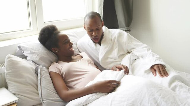 Happy black man surprisingly looks at pregnant wife's belly, man is in shock, every day baby in stomach becomes bigger and bigger, they lie on bed together.