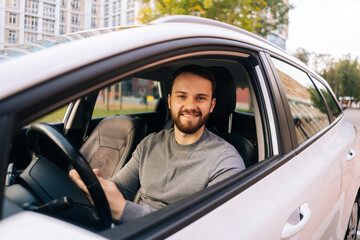 Cheerful young man sitting in car and using phone in auto, looking at camera. Male sitting in vehicle and works on smartphone.