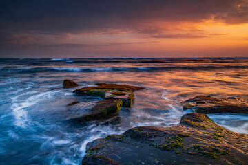 Beautiful seascape. Beach with stones covered by seaweeds. Low tide. Composition of nature. Motion water. Cloudy sky with sunlight. Slow shutter speed. Mengening beach, Bali, Indonesia