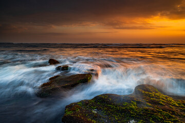 Obraz premium Beautiful seascape. Beach with stones covered by seaweeds. Low tide. Composition of nature. Motion water. Cloudy sky with sunlight. Slow shutter speed. Mengening beach, Bali, Indonesia