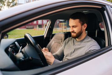 Caucasian bearded young man sitting in car and typing online message on cell phone, side view. Handsome businessman using mobile phone in auto. Male sitting in modern vehicle and works on smartphone.
