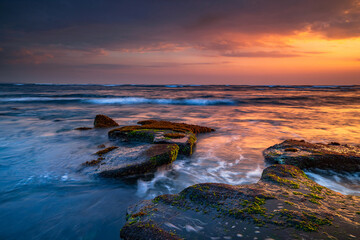 Obraz premium Beautiful seascape. Beach with stones covered by seaweeds. Low tide. Composition of nature. Motion water. Cloudy sky with sunlight. Slow shutter speed. Mengening beach, Bali, Indonesia