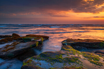 Beautiful seascape. Beach with stones covered by seaweeds. Low tide. Composition of nature. Motion water. Cloudy sky with sunlight. Slow shutter speed. Mengening beach, Bali, Indonesia