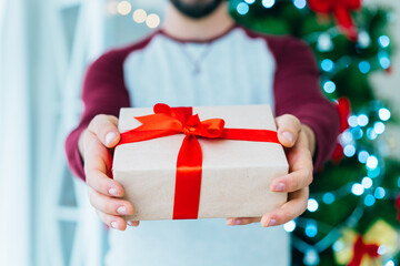 Close up cropped shot of man hands holding gift box with the Christmas tree on the background