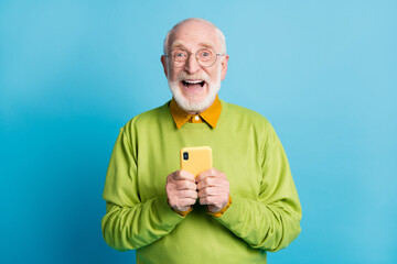 Photo of pensioner old man hold smartphone excited face wear glasses green sweater isolated blue...
