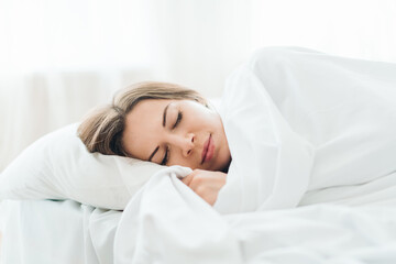 Portrait of a young woman sleeping in cozy light colored bedroom lying in the bed with eyes closed