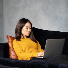 Asian woman looking at laptop, working at home, liying on sofa. Unhappy, upset Japanese female freelancer smiling and typing at computer in hotel on couch