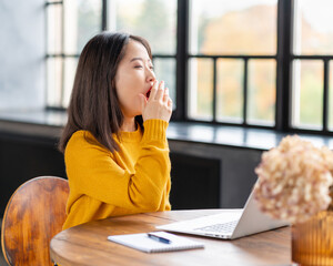 Asian woman yawning due to overworking and exhausting. Young lady in bright yellow jumper working on laptop at home or in cafe. Business oriental female