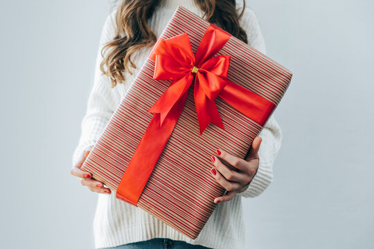 Cropped Shot Of A Girl In White Knitted Oversized Sweater Holding Big Gift Box Decorated With Beautiful Red Satin Ribbon Bow