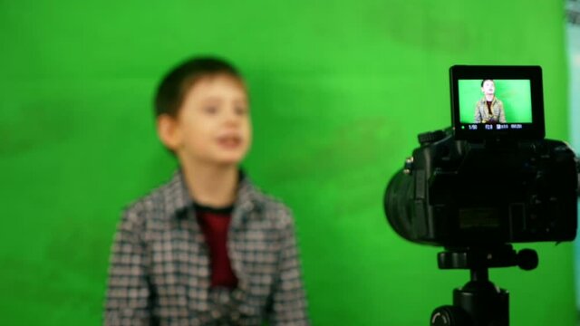 Cute caucasian preschooler boy writes a blog while sitting in front of a video camera on a background of green cloth. The kid keeps his blog. Little blogger. Selective focus, child out of focus