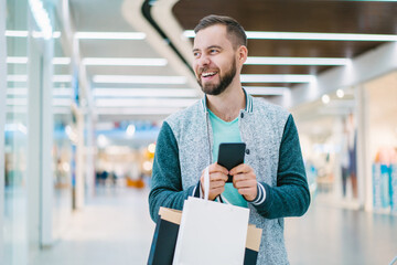 Handsome smiling young bearded man in casual outfit looking at the show-window while walking in a...