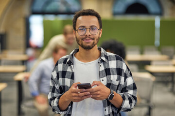 Fototapeta na wymiar Portrait of a young happy mixed race man holding smartphone and looking at camera while working in the modern office, selective focus