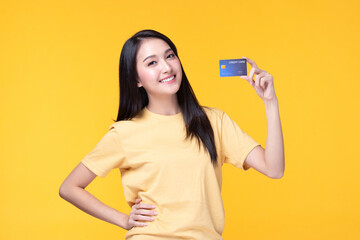 Cheerful young asian woman in yellow T-shirt holding bank card isolated on yellow background