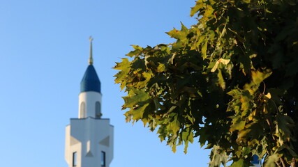 abstract background symbolizing the faith of Islam, green maple foliage in the city park and the blue and white tower of the mosque in the blurred background