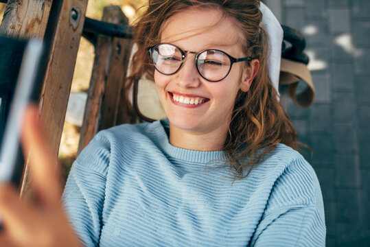 Positive female in the a blue sweater, eyeglasses, smiling and lying on the bench taking selfie on a mobile phone. Happy woman takes a rest during an online conversation on a smartphone outside