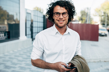 Confident businessman in eyewear smiling broadly posing outdoors. Male entrepreneur messaging on smartphone. Smart guy in casual wears spectacles with curly hair walking outside after work
