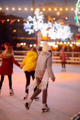 Young woman ice skating on a rink in a Festive Christmas fair in the evening. Smiling woman in winter style clothes skates.  Winter holidays concept. Lights around. - obrazy, fototapety, plakaty