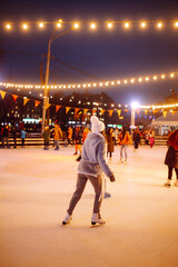 Young woman ice skating on a rink in a Festive Christmas fair in the evening. Smiling woman in winter style clothes skates.  Winter holidays concept. Lights around.