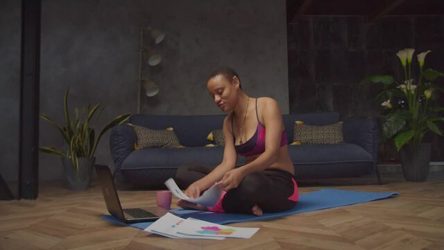 Attractive sporty african businesswoman sitting cross legged on exercise mat, video conferencing online on laptop with partner, discussing financial charts and graphs while working after training.
