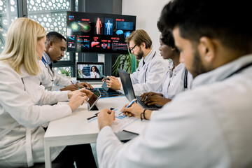 Group of multiethnical healthcare workers with digital tablets and laptop, meeting in hospital boardroom. Medical staff during morning online briefing with their chief, African lady, with CT scan