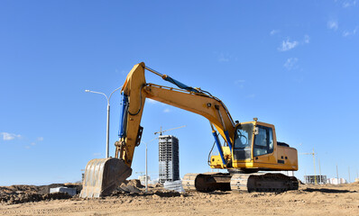 View of a large construction site where earthmoving equipment. Excavator digs the ground to lay pipes and a construction new road. Tower cranes are building tall residential buildings.