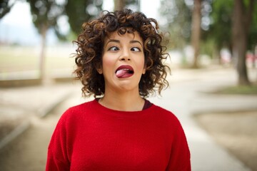 Young arab woman wearing casual red sweater in the street, showing grimace face crossing her eyes and showing tongue. Being funny and crazy - obrazy, fototapety, plakaty