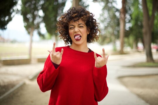 Young arab woman wearing casual red sweater in the street, making rock hand gesture and showing tongue