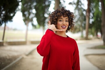 Young arab woman wearing casual red sweater in the street, smiling doing phone gesture with hand...