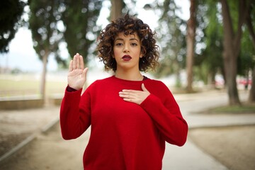 Young arab woman wearing casual red sweater in the street, Swearing with hand on chest and open...