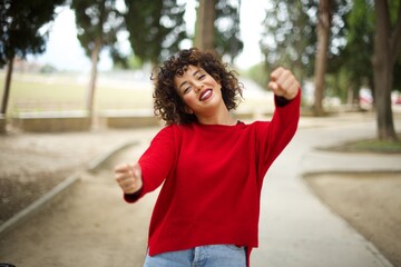 Young arab woman wearing casual red sweater in the street, imagine steering wheel helm rudder passing driving exam good mood fast speed
