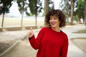 Young beautiful Arab woman standing outdoors wearing red sweater Looking proud, smiling doing thumbs up gesture to the side. Good job!