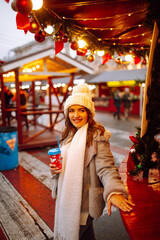 Young woman drinking hot coffee while walking in Christmas market decorated with holiday lights in the evening. Christmas and New Year magic. Lights around.