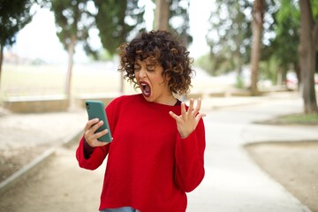 Angry Young beautiful Arab woman standing outdoors wearing red sweater screaming on the phone,...