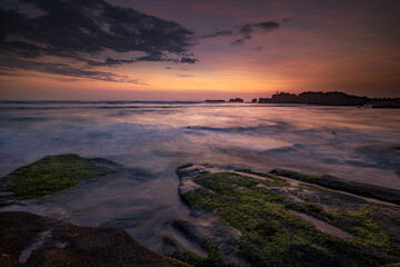 Fototapeta na wymiar Calm ocean long exposure. Stones covered by green moss in mysterious mist of the sea waves. Concept of nature background. Sunset scenery background. Mengening beach, Bali, Indonesia.