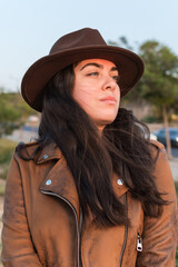 Young woman in hat and brown leather jacket, looking into the distance, in natural park.