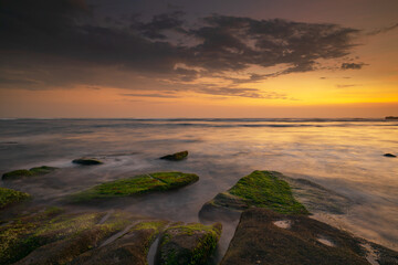 Calm ocean long exposure. Stones covered by green moss in mysterious mist of the sea waves. Concept of nature background. Sunset scenery background. Mengening beach, Bali, Indonesia.