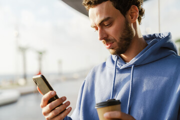 Smiling young guy drinking coffee and using cellphone