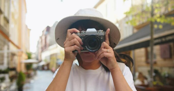 Close up portrait of cheerful Asian beautiful female in hat and glasses taking photo on camera while standing on street. Joyful pretty woman tourist takes pictures outdoors. Urban tourism concept
