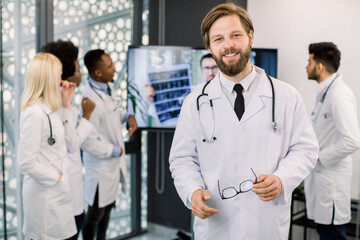 Confident smiling young doctor posing with eyeglasses to camera at the hospital. International medical team working on the background, analyzing patient's CT, consulting with Caucasian doctor online