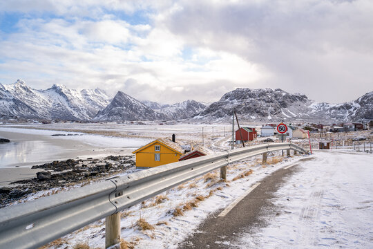 Snowy empty road along the sea & shoreline & beach in Lofoten. Yellow and red houses are on the side of the street. Road barrier / guard rails for vehicles