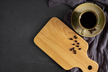 Top view above of Black hot fresh coffee and wooden chopping board with handle and hole for hanging and coffee beans roasted, apron on dark black table background. with copy space.